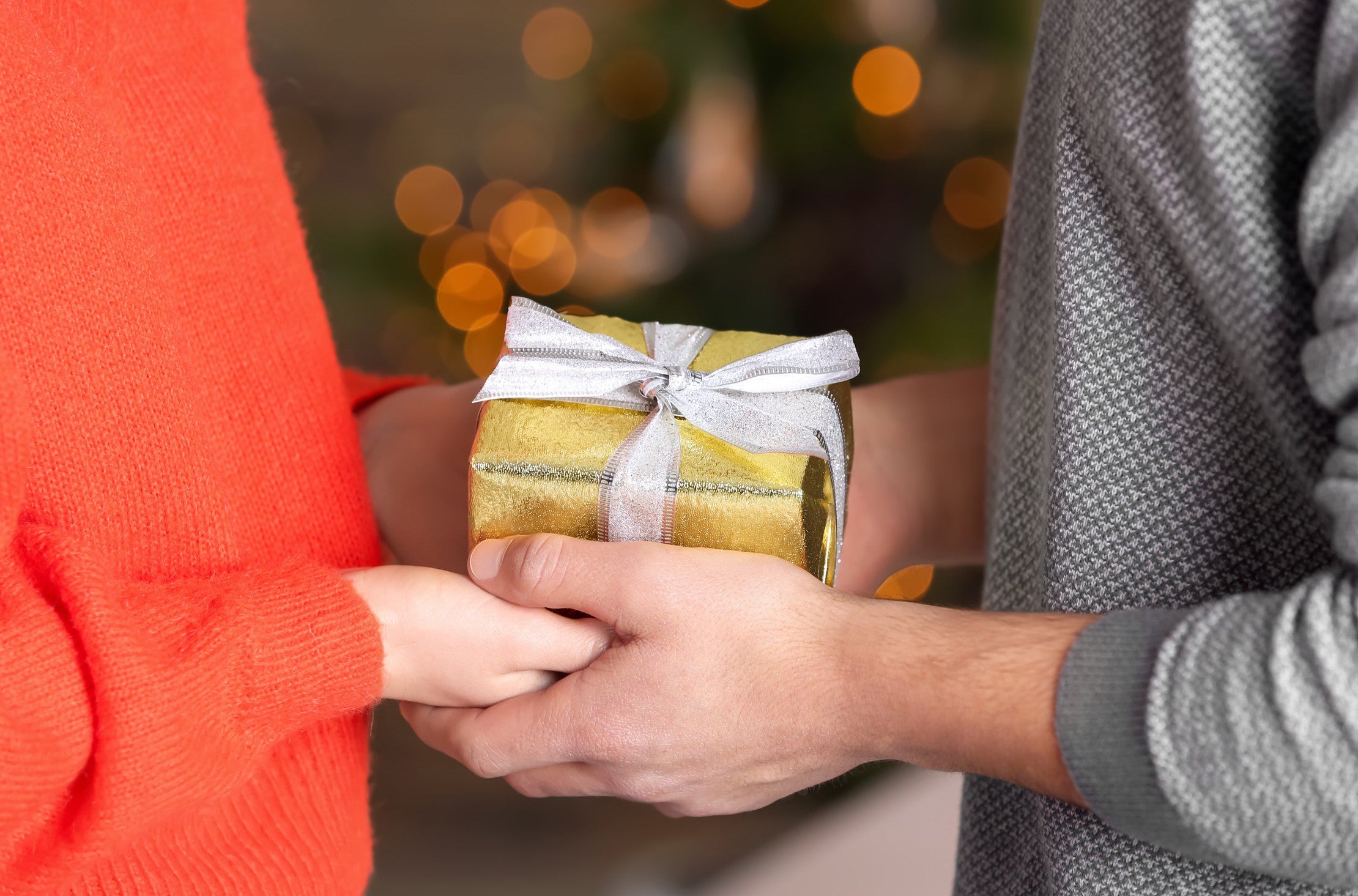 Two people exchanging a gold gift box with a white ribbon against a blurred festive background.